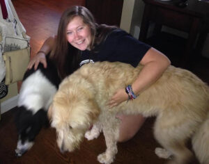 NECC student Olivia Lucey is crouched on a hardwood floor holding her two large breed dogs, Buddy and Maisy.