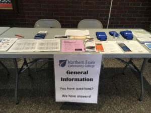A plastic foldable table with prices of paper and other items on it. The stable has a pice of paper taped to it that has necc's logo and reads general information. 