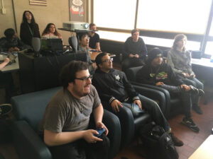 Several people siting holding controllers. Thare is a group in the front citing on two couches, and another group in the back standing 
