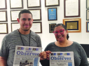 Man and woman standing next to eachother holding print copies of the NECC Observer with awards plaques in the background. 