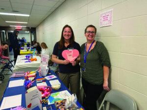 Two women standing by a table in a hall, one of them holding a cutout paper heart. 