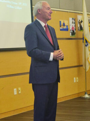 A man in a suit standing in front of a room speaking. 
