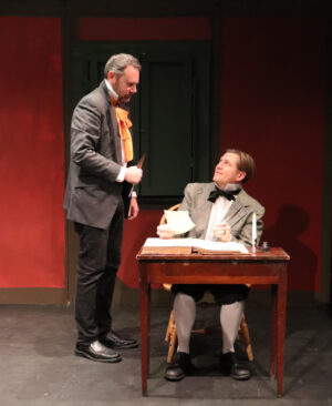 A standing man talks to a man sitting at a desk in a scene from "A Christmas Carol."