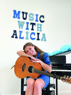 A woman sitting at a piano holding a guitar with a sign in the background advertising Music with a Alicia