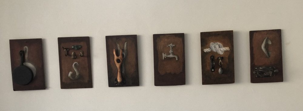 A series of artwork hanging on wall. Drawings or paintings of various objects. A faucet, a pan, and something that appears to be hooks and tongs.