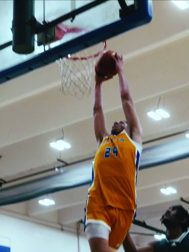 A male basketball player leaps into the air to attempt to put the ball in the basket while another player tries to block the shot.