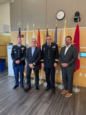 Four men, including two in military uniforms, stand in front of flags in the Tech Center building.