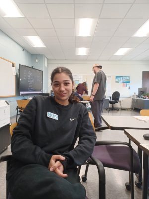 A woman sits in a chair in a classroom and looks at the camera.