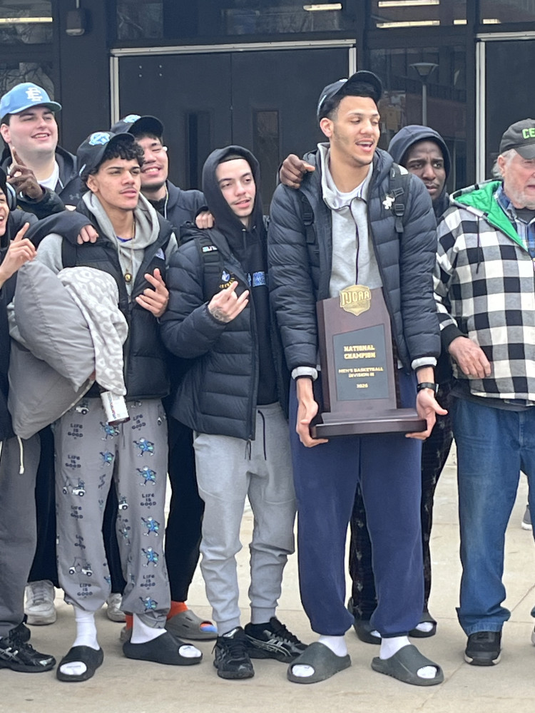 Members of the basketball team stand in a line holding their national championship trophy.