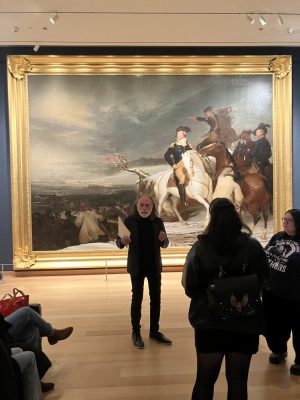 A man stands in front of a large painting and talks about it to a crowd at the Museum of Fine Arts in Boston. 