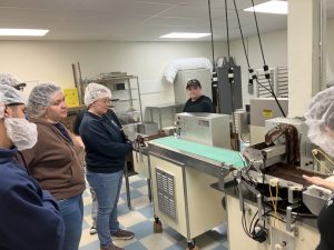 PACE TRIO staff on Van Otis chocolate factory tour. Group of men and women watch chocolate being made on a conveyor belt.